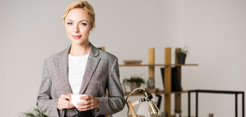 Donna d’affari bionda in piedi nel suo ufficio con una tazza di tè tra le mani che guarda la fotocamera